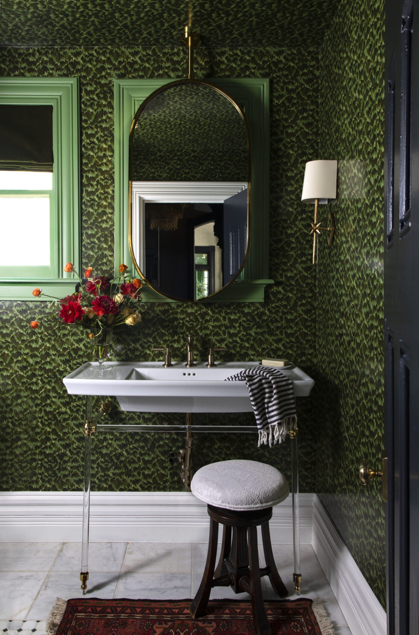 Rivington Marx Interiors – Green Wall Decor Denver powder room with oval brass-framed mirror over a white sink with clear acrylic legs and a dark wood stool