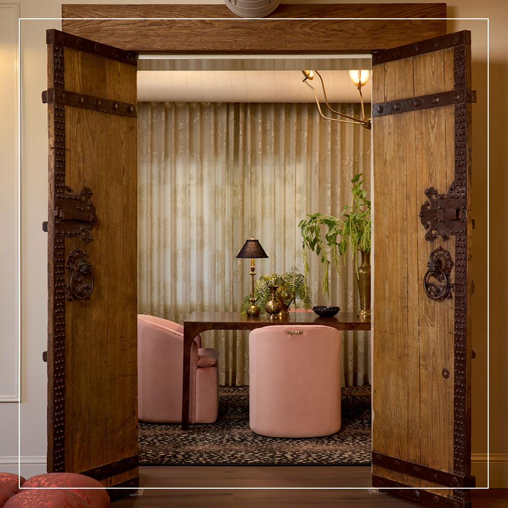 View through wood doors to small area with pink velvet chairs, a table with a lamp and vases, and tall curtains