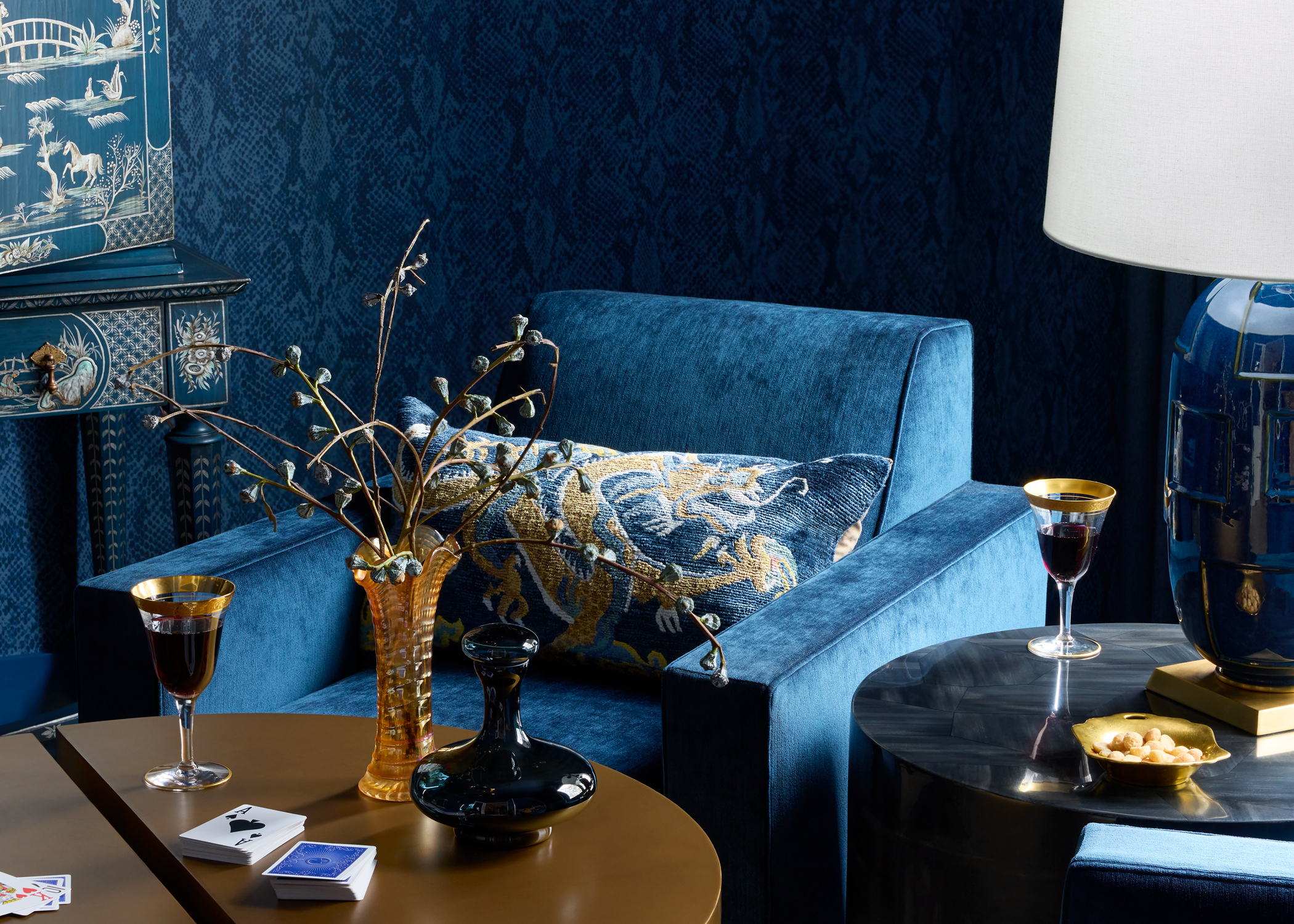 Elegant living room corner with a rich blue armchair, gold-patterned pillow, and chic decor. Nearby are a lamp, two wine glasses, and small tables.