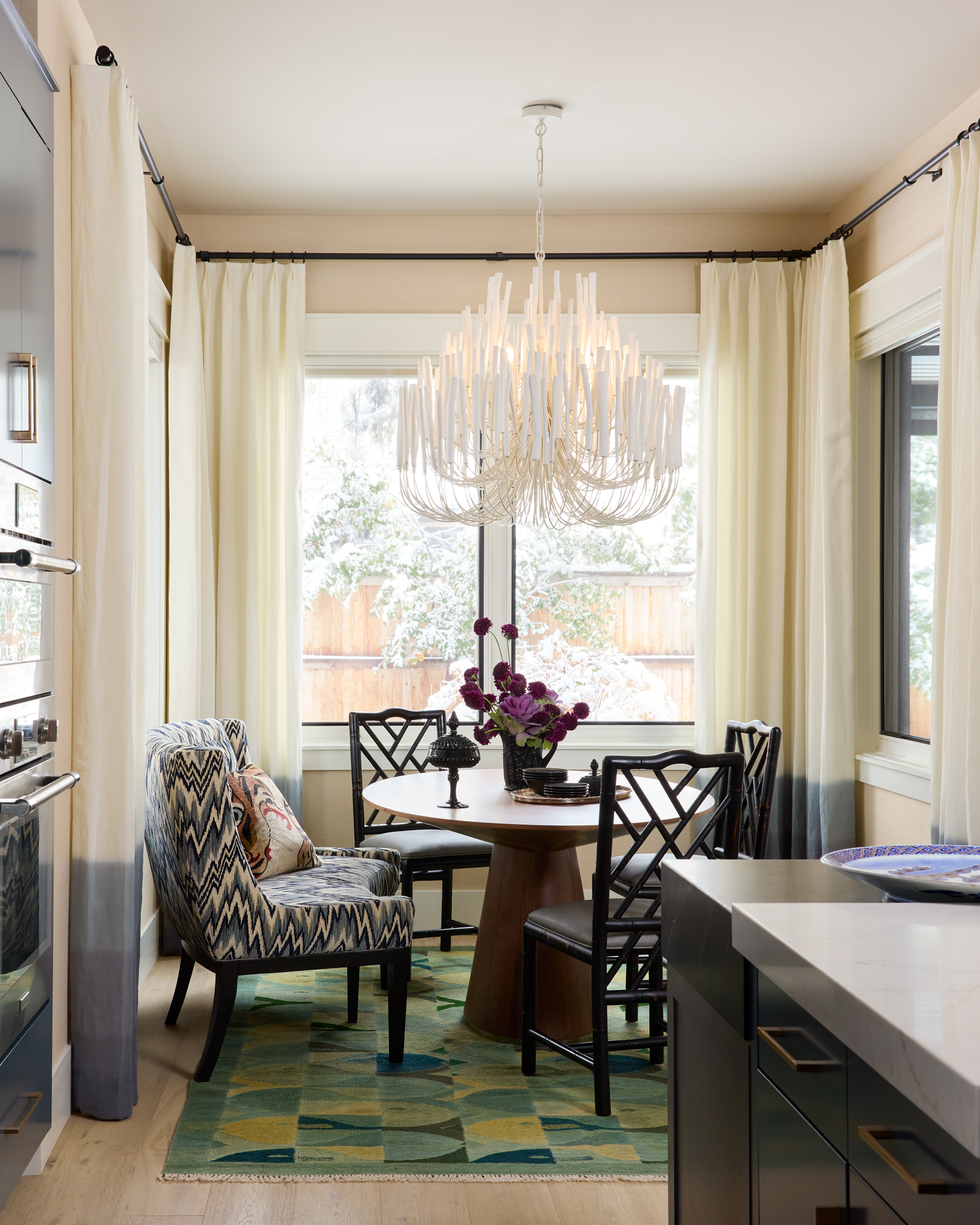 Stylish dining room with a round table, eclectic chairs, and a chic chandelier. Large windows with drapes reveal a snowy yard, adding warmth and elegance.