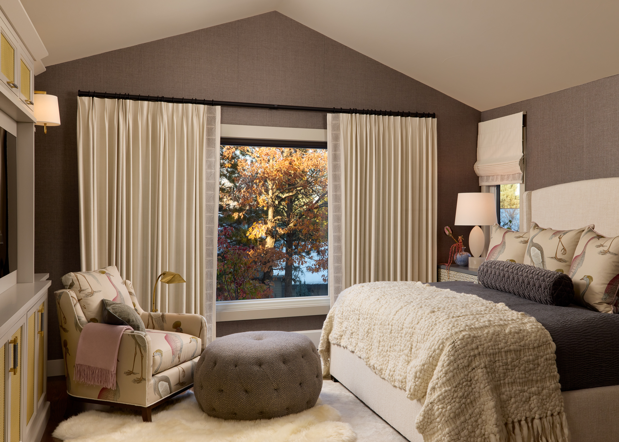 Cozy bedroom with soft lighting and autumn view. Features a plush bed, armchair, ottoman, and large window. Warm, inviting atmosphere.