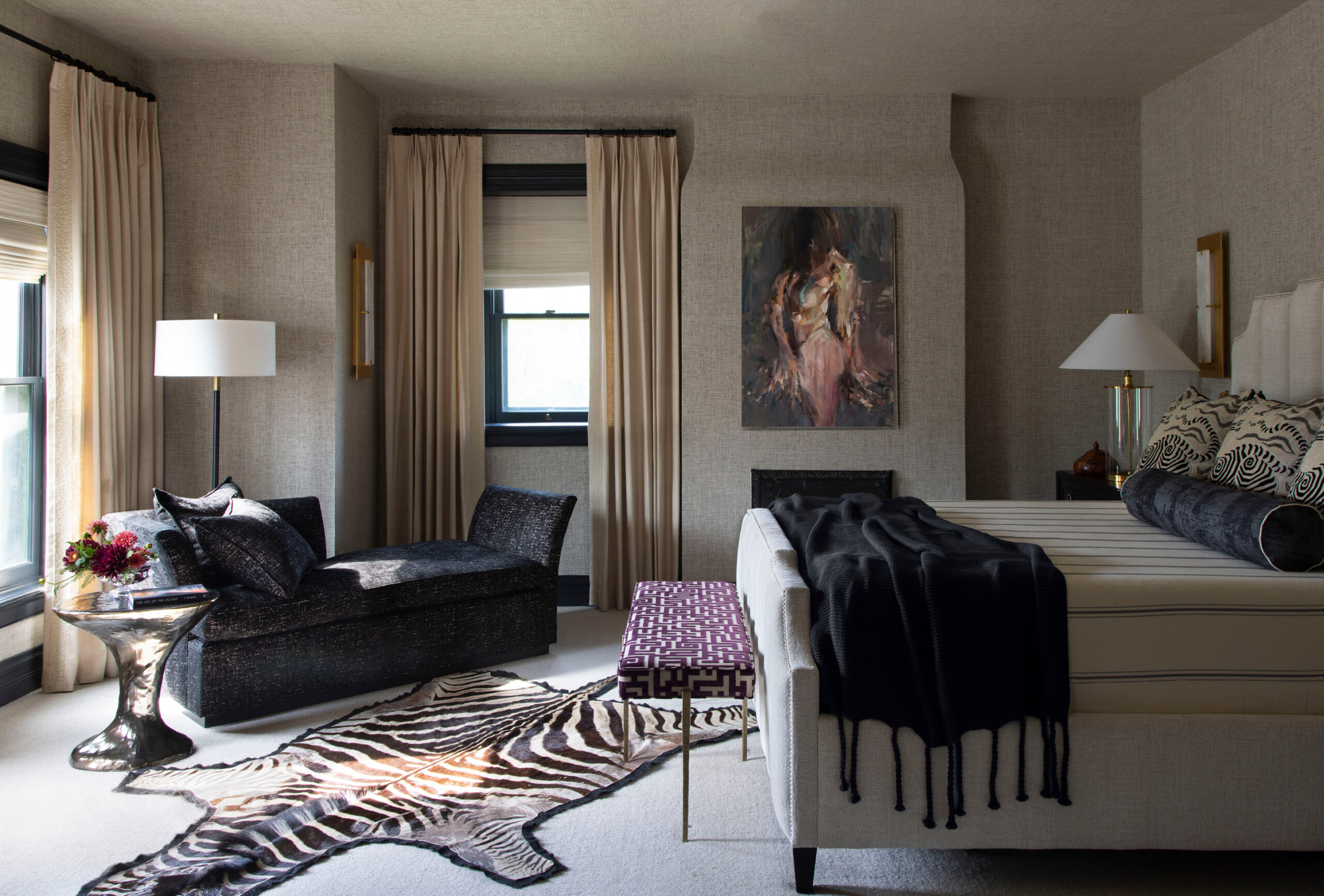 Stylish bedroom with neutral tones, featuring a zebra print rug, black chaise, patterned ottoman, abstract artwork, and large windows with beige curtains.